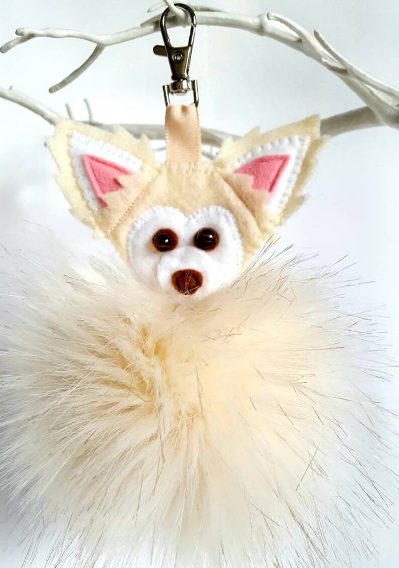 A white faux fur pom pom keychain with a felt chihuahua head attached. The chihuahua has pink ears and brown bead eyes.