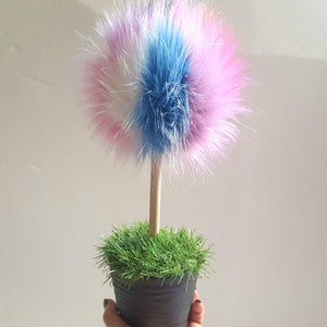 Puede incluir: Un &aacute;rbol de pomp&oacute;n esponjoso y colorido en una maceta negra peque&ntilde;a con hierba verde. El pomp&oacute;n est&aacute; hecho de piel rosa, blanca y azul.