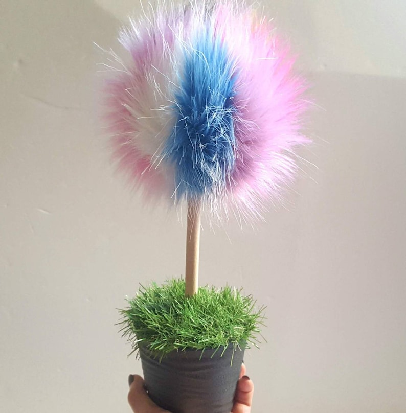 Puede incluir: Un &aacute;rbol de pomp&oacute;n esponjoso y colorido en una maceta negra peque&ntilde;a con hierba verde. El pomp&oacute;n est&aacute; hecho de piel rosa, blanca y azul.