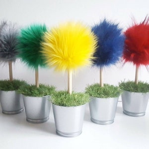 May include: Five decorative topiary-style ornaments featuring fluffy, spherical faux fur pom-poms in gray, green, yellow, blue, and red. Each pom-pom is attached to a light-colored wooden dowel stem, set into a silver metal bucket filled with green grass.