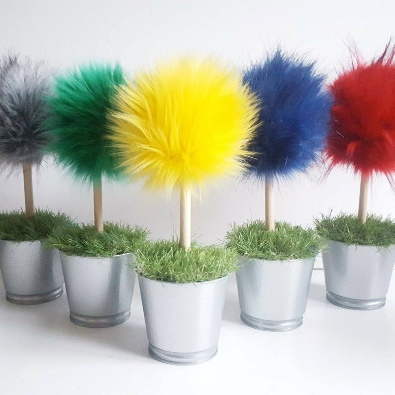 May include: Five decorative topiary-style ornaments featuring fluffy, spherical faux fur pom-poms in gray, green, yellow, blue, and red. Each pom-pom is attached to a light-colored wooden dowel stem, set into a silver metal bucket filled with green grass.