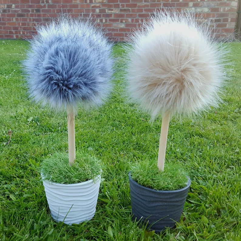 May include: Two decorative topiary ornaments featuring fluffy, spherical faux fur pom-poms in grey and beige. Each pom-pom is mounted on a straight, light-colored wooden dowel stem, which is set into a small, textured pot filled with green faux grass.