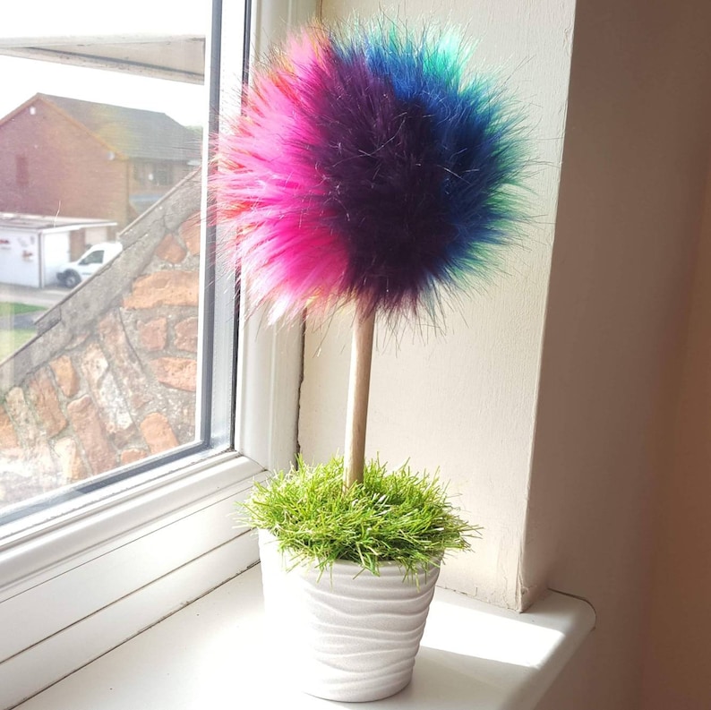 Puede incluir: Un &aacute;rbol de pompones de pelo sint&eacute;tico de colores en una maceta blanca con hierba verde. El pompon es rosa, morado, azul y verde.