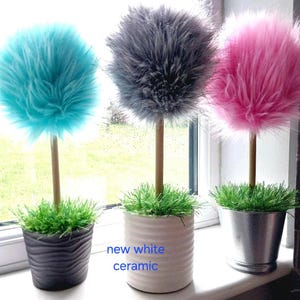 May include: Three decorative topiary plants with fluffy, spherical faux fur foliage in turquoise, gray, and pink. Each plant has a thin wooden stem, a base of green artificial grass, and sits in a pot: black wavy, new white ceramic, or silver metal.