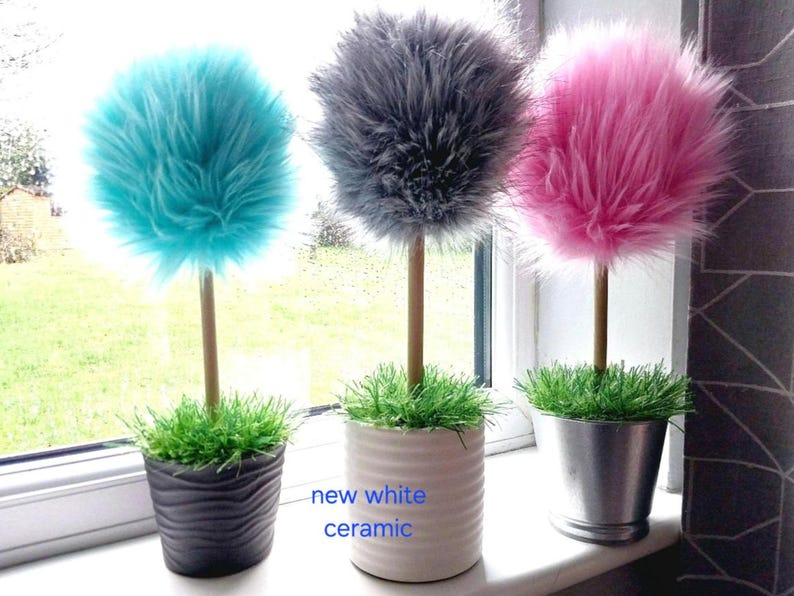 May include: Three decorative topiary plants with fluffy, spherical faux fur foliage in turquoise, gray, and pink. Each plant has a thin wooden stem, a base of green artificial grass, and sits in a pot: black wavy, new white ceramic, or silver metal.