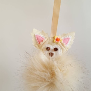 May include: A white felt chihuahua ornament with a pink flower on its head, hanging from a beige ribbon. The ornament is attached to a fluffy white pom-pom.