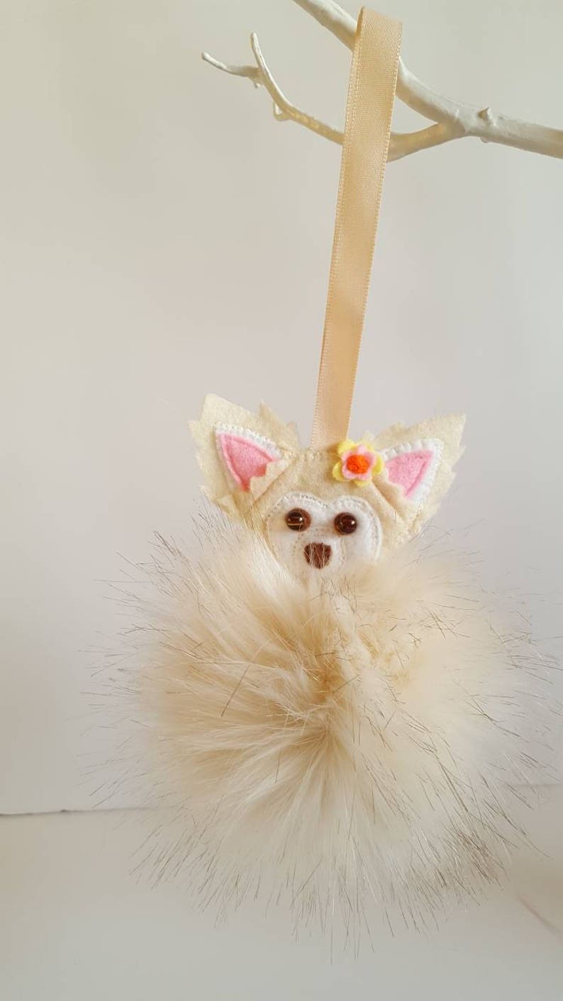 May include: A white felt chihuahua ornament with a pink flower on its head, hanging from a beige ribbon. The ornament is attached to a fluffy white pom-pom.
