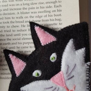 Op de afbeelding: Een zwart en wit vilt kattenbladwijzer met roze oren en neus. De kat heeft groene ogen en een witte vlek op zijn gezicht.