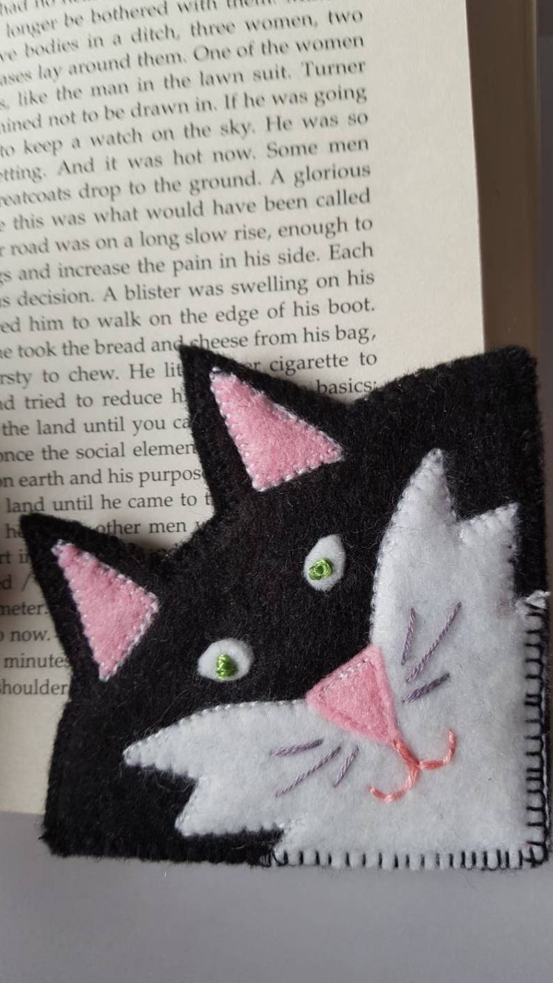 Op de afbeelding: Een zwart en wit vilt kattenbladwijzer met roze oren en neus. De kat heeft groene ogen en een witte vlek op zijn gezicht.