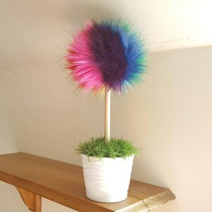 Puede incluir: Un pomp&oacute;n de pelo sint&eacute;tico arco&iacute;ris en un palo de madera en una maceta blanca con hierba verde.