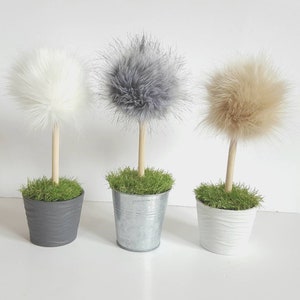 May include: Three decorative topiary ornaments featuring fluffy faux fur pom-poms in white, gray, and beige. Each pom-pom is attached to a light-colored wooden dowel stem, set into a small pot filled with artificial green grass.