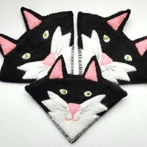 Op de afbeelding: Drie zwarte en witte vilt kattengezichtsversieringen met roze oren en groene ogen. De kattengezichten zijn in een driehoekige vorm gerangschikt.