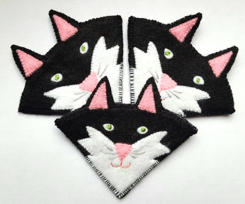 Op de afbeelding: Drie zwarte en witte vilt kattengezichtsversieringen met roze oren en groene ogen. De kattengezichten zijn in een driehoekige vorm gerangschikt.