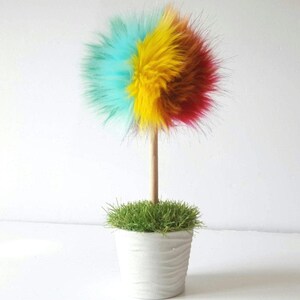 Puede incluir: Un &aacute;rbol de pomp&oacute;n de piel sint&eacute;tica de colores en una maceta blanca con hierba verde. El pomp&oacute;n est&aacute; hecho de piel sint&eacute;tica turquesa, amarilla y roja.