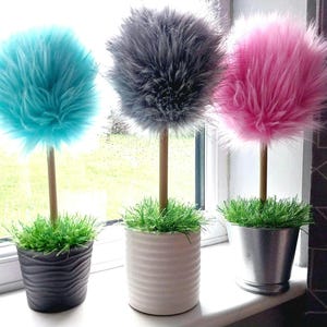 May include: Three decorative topiary-style ornaments featuring fluffy, spherical faux-fur pom-poms in bright turquoise, dark grey, and vibrant pink. Each pom-pom sits atop a thin wooden dowel stem, anchored in a pot filled with green faux grass.