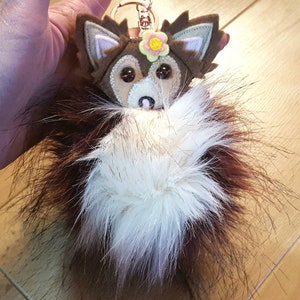 May include: A brown and white furry keychain with a felt fox head and a yellow flower. The fox has brown ears, a white face, and brown eyes.