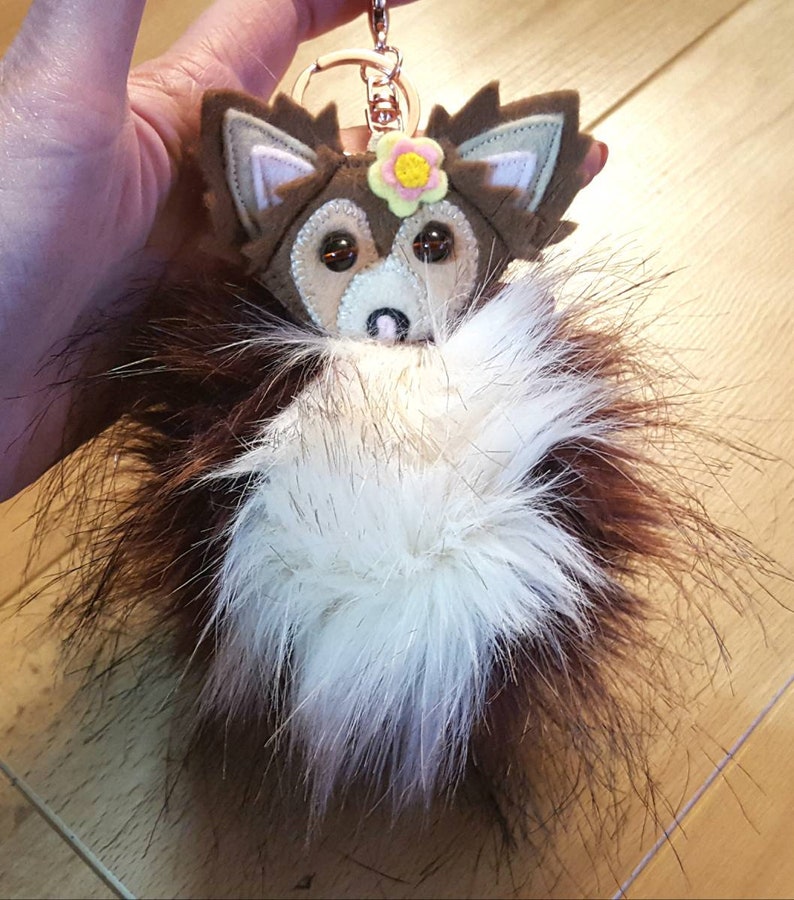 May include: A brown and white furry keychain with a felt fox head and a yellow flower. The fox has brown ears, a white face, and brown eyes.