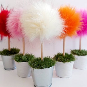 May include: Five decorative topiary-style ornaments featuring fluffy, round faux fur pom-poms in red, light pink, white, orange, and bright pink. Each pom-pom sits atop a light wooden dowel stem, planted in a small silver or white metal bucket.