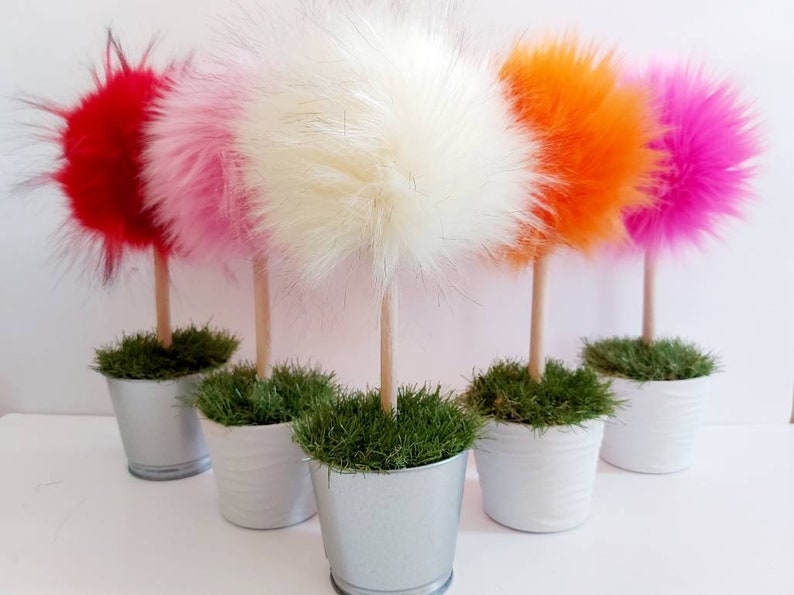 May include: Five decorative topiary-style ornaments featuring fluffy, round faux fur pom-poms in red, light pink, white, orange, and bright pink. Each pom-pom sits atop a light wooden dowel stem, planted in a small silver or white metal bucket.
