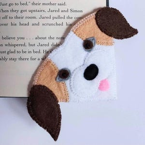 May include: A felt corner bookmark shaped like a dog head with white, tan, and dark brown patches. The dog features two dark brown ears, two dark eyes, a black nose, and a small pink tongue, all assembled with visible light-colored stitching.