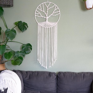 May include: A white macrame wall hanging in the shape of a tree with a circular frame. The wall hanging is on a green wall with a gray couch and a potted plant in front of it.