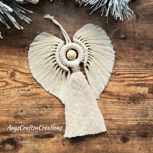 May include: Cream-colored macrame angel ornament with a wooden bead head and a long tassel skirt. The wings are fanned out, and the ornament is set against a wooden background with frosted pine and red berries.