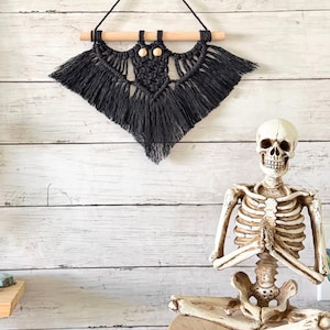 May include: A black macrame wall hanging shaped like a bat, featuring two light-colored wooden beads for eyes, suspended from a horizontal wooden dowel with a black cord loop for hanging, and a full-body human skeleton model in a seated pose.