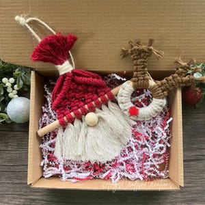 May include: A set of two handmade Christmas ornaments in a cardboard box. The ornaments are made of yarn and feature a Santa Claus and a reindeer. The ornaments are red, white, and brown.