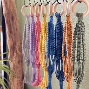 May include: A set of eight macrame plant hangers in various colors, including gray, purple, pink, yellow, blue, and green. Each hanger is made with a wooden ring and has a tassel at the bottom.