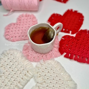 Puede incluir: Una taza de té se encuentra sobre un posavasos de macramé rosa, rodeada de otros posavasos en forma de corazón en rosa, rojo y blanco. Un carrete de hilo rosa y un rollo de hilo rojo están en el fondo. El texto "AngsCraftsnCreations" es visible.