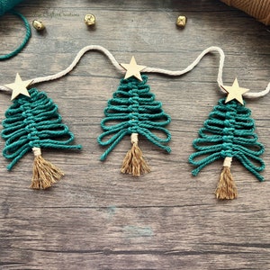 May include: A garland of three green macrame Christmas trees with gold stars and tassels hanging on a white string against a wood background.