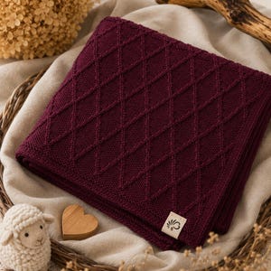 Handgestrickte Babydecke aus mulesingfreier Merinowolle in Bordeaux, dekorativ auf Leinenstoff mit Holzherz, Schäfchen und Trockenblumen arrangiert – nachhaltige Babydecke für Neugeborene und stilvolles Geschenk zur Geburt.