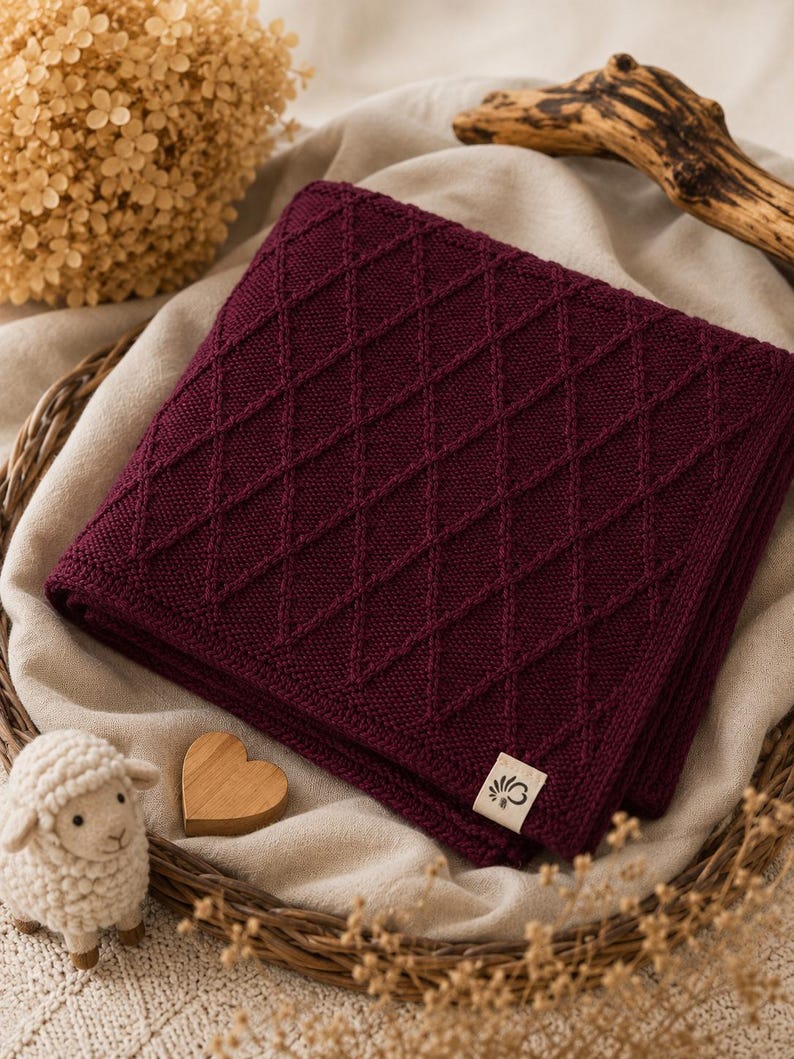 Handgestrickte Babydecke aus mulesingfreier Merinowolle in Bordeaux, dekorativ auf Leinenstoff mit Holzherz, Schäfchen und Trockenblumen arrangiert – nachhaltige Babydecke für Neugeborene und stilvolles Geschenk zur Geburt.