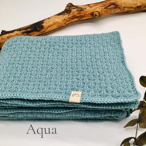 Handgestrickte Babydecke in der Farbe Aqua mit feiner Strickstruktur, weiches Naturgarn mit Joseda-Label, dekoriert mit Holzast und Blättern auf hellem Hintergrund, Farbbezeichnung „Aqua“ im Bild.