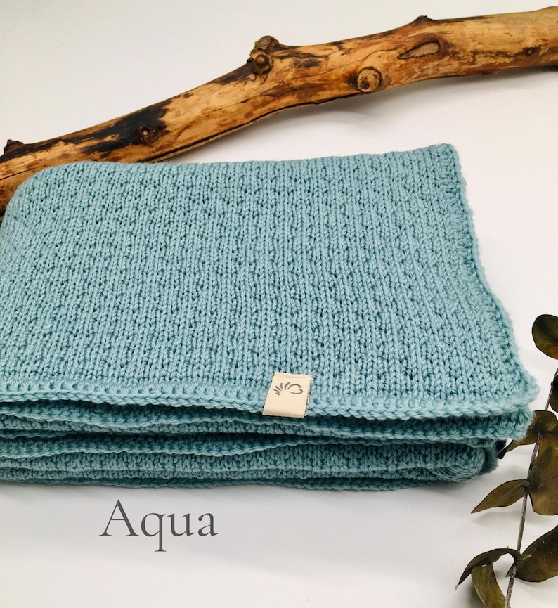 Handgestrickte Babydecke in der Farbe Aqua mit feiner Strickstruktur, weiches Naturgarn mit Joseda-Label, dekoriert mit Holzast und Blättern auf hellem Hintergrund, Farbbezeichnung „Aqua“ im Bild.