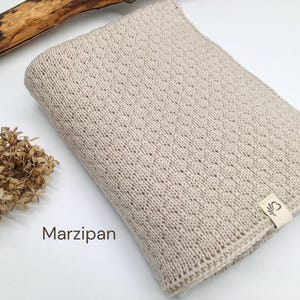 Handgestrickte Babydecke in der Farbe Marzipan, weiche Baumwoll-Strickdecke mit feiner Struktur, Joseda-Label, dekoriert mit Trockenblumen und Holzast auf weißem Hintergrund.