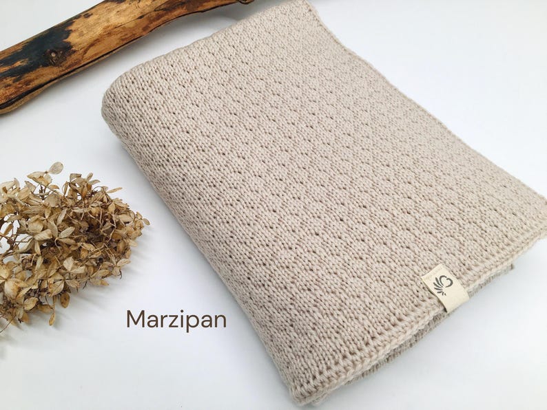 Handgestrickte Babydecke in der Farbe Marzipan, weiche Baumwoll-Strickdecke mit feiner Struktur, Joseda-Label, dekoriert mit Trockenblumen und Holzast auf weißem Hintergrund.