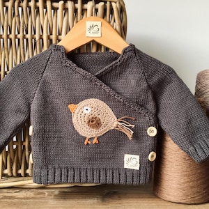 Op de afbeelding: Een donkergrijze babyvest met een overslagvoorkant en houten knopen. De vest heeft een gehaakte vogelapplicatie in beige, bruin en oranje. De vest wordt getoond op een houten hanger voor een rieten mand.