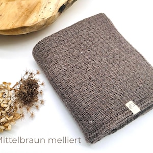 Handgestrickte Babydecke in Mittelbraun meliert, weiche Strickdecke mit feiner Struktur, hochwertiges Naturgarn, Joseda-Label, dekoriert mit Trockenblumen auf weißem Hintergrund.