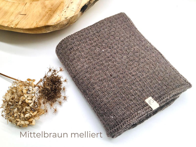 Handgestrickte Babydecke in Mittelbraun meliert, weiche Strickdecke mit feiner Struktur, hochwertiges Naturgarn, Joseda-Label, dekoriert mit Trockenblumen auf weißem Hintergrund.