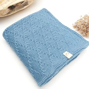 May include: A folded light blue knitted blanket featuring a textured diamond pattern across the surface. A small rectangular beige fabric tag with a simple line drawing logo is attached to the bottom right corner of the knitted fabric.