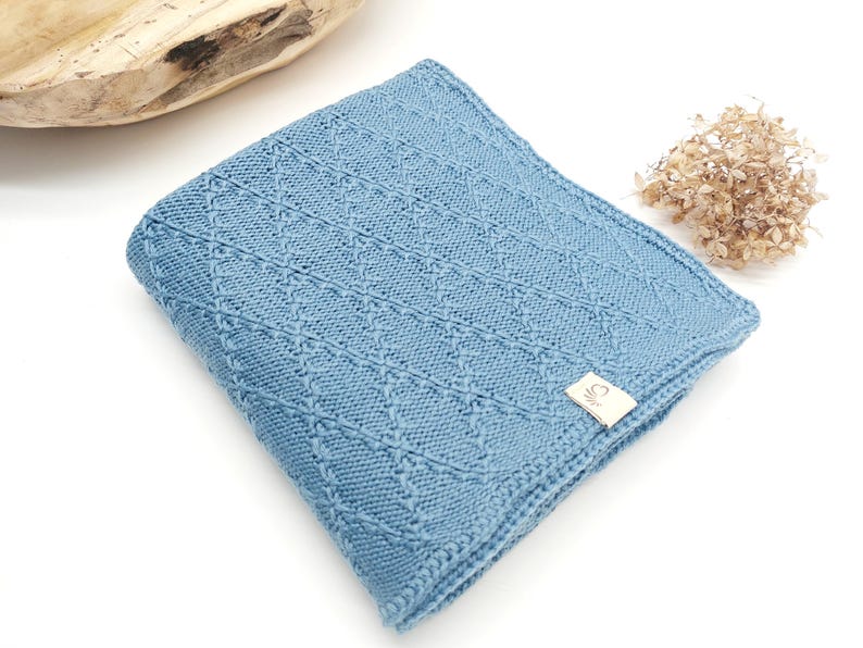 May include: A folded light blue knitted blanket featuring a textured diamond pattern across the surface. A small rectangular beige fabric tag with a simple line drawing logo is attached to the bottom right corner of the knitted fabric.
