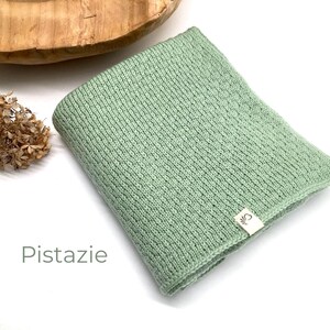 Handgestrickte Babydecke in der Farbe Pistazie mit feiner Strickstruktur, weiches Naturgarn mit Joseda-Label, dekoriert mit Trockenblumen und Holzschale auf hellem Hintergrund, Farbbezeichnung „Pistazie“ im Bild.