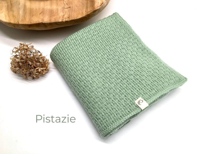 Handgestrickte Babydecke in der Farbe Pistazie mit feiner Strickstruktur, weiches Naturgarn mit Joseda-Label, dekoriert mit Trockenblumen und Holzschale auf hellem Hintergrund, Farbbezeichnung „Pistazie“ im Bild.