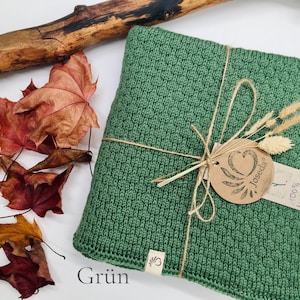 Handgestrickte Decke in Grün, mit Naturkordel gebunden und Joseda-Etikett, dekoriert mit Herbstblättern und Holzast auf hellem Hintergrund.