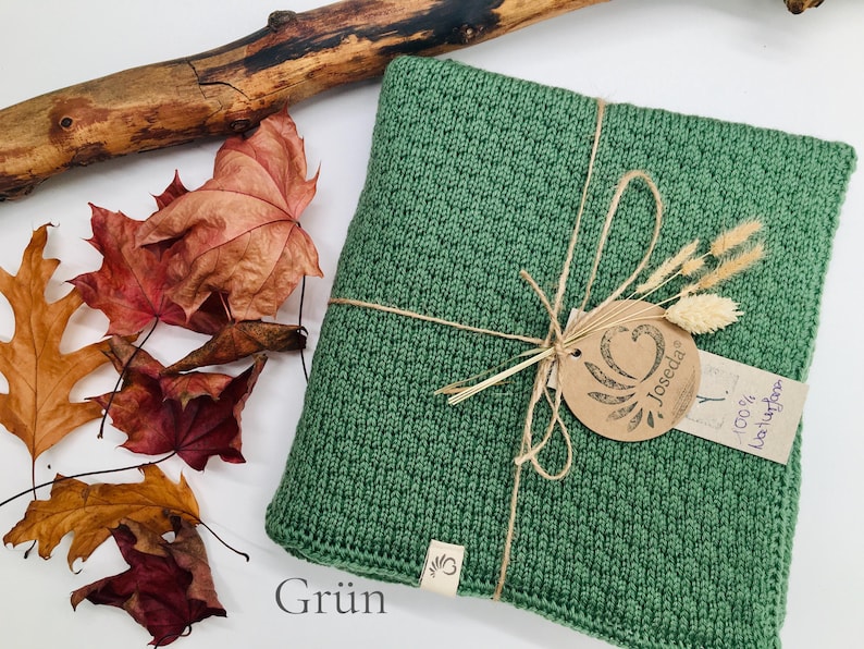 Handgestrickte Decke in Grün, mit Naturkordel gebunden und Joseda-Etikett, dekoriert mit Herbstblättern und Holzast auf hellem Hintergrund.