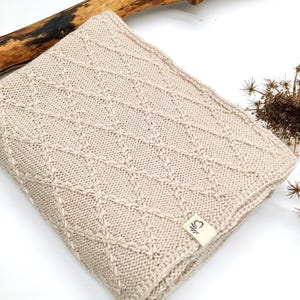 May include: A folded beige knit blanket featuring a raised diamond pattern across the surface. A small rectangular fabric tag with a black graphic logo is attached to the bottom corner of the blanket, which is made of a textured yarn material.