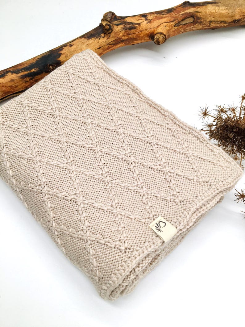 May include: A folded beige knit blanket featuring a raised diamond pattern across the surface. A small rectangular fabric tag with a black graphic logo is attached to the bottom corner of the blanket, which is made of a textured yarn material.