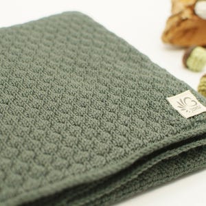 Könnte beinhalten: Eine gefaltete, olivgrüne Strickdecke mit strukturiertem Muster. Ein kleines Etikett ist an der Decke befestigt. Dekorative Eicheln befinden sich im Hintergrund.