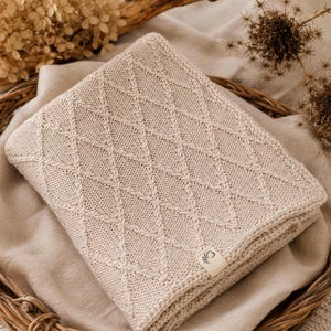 Handgestrickte Babydecke aus mulesingfreier Merinowolle in Naturbeige, dekorativ auf einem Leinentuch mit Holz, Trockenblumen und Herz in Szene gesetzt – nachhaltige Babydecke für Neugeborene und Geschenk zur Geburt.
